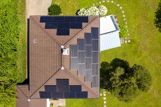Senkrechte Luftaufnahme eines Einfamilienhauses mit braunem Ziegeldach; auf mehreren Dachflächen sind schwarze Photovoltaikmodule montiert. Das Haus ist von einem grünen Garten mit Rasen, Bäumen und einer gepflasterten Wegführung umgeben.